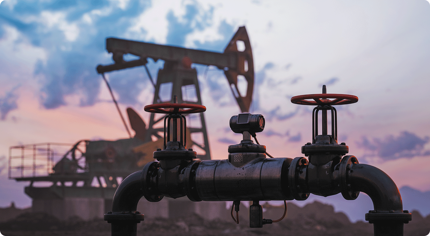 A close-up of an oil pipeline valve with a pumpjack in the background at sunset.